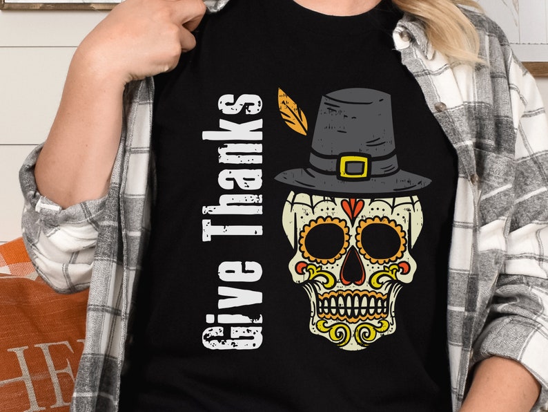 May include: A black t-shirt featuring a graphic of a sugar skull wearing a pilgrim hat with a yellow feather. The text "Give Thanks" is printed vertically in white distressed lettering to the left of the skull, which has orange and yellow accents.