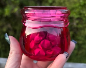 May include: A small, translucent, hot pink glass jar with a rounded body and a threaded rim. The interior of the jar is filled with multiple small, folded, dark pink paper stars, creating a dense collection of geometric shapes inside the container.
