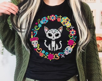 May include: A black t-shirt featuring a central graphic of a cat with sugar skull face markings, surrounded by a colorful circular wreath of flowers, hearts, candles, and multiple sugar skulls in shades of pink, yellow, blue, and red.