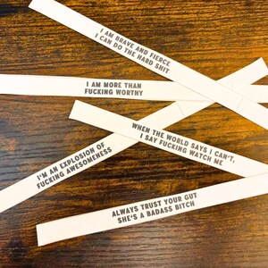 May include: Five white paper strips featuring black printed text with motivational phrases: I AM BRAVE AND FIERCE I CAN DO THE HARD SHIT, I AM MORE THAN FUCKING WORTHY, WHEN THE WORLD SAYS I CAN'T, I SAY FUCKING WATCH ME, I'M AN EXPLOSION OF FUCKING AWESOMENESS, and ALWAYS TRUST YOUR GUT SHE'S A BADASS BITCH.