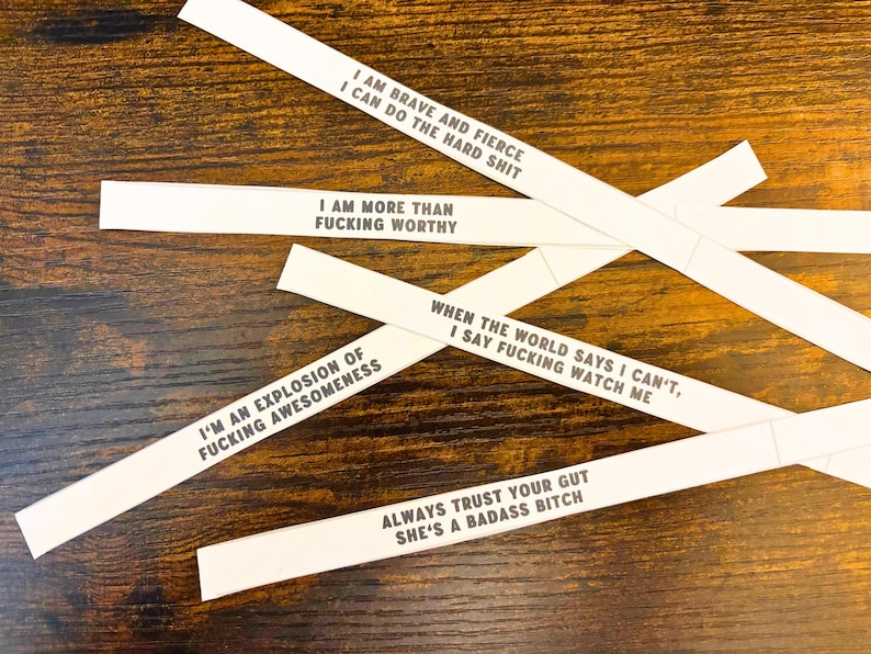 May include: Five white paper strips featuring black printed text with motivational phrases: I AM BRAVE AND FIERCE I CAN DO THE HARD SHIT, I AM MORE THAN FUCKING WORTHY, WHEN THE WORLD SAYS I CAN'T, I SAY FUCKING WATCH ME, I'M AN EXPLOSION OF FUCKING AWESOMENESS, and ALWAYS TRUST YOUR GUT SHE'S A BADASS BITCH.