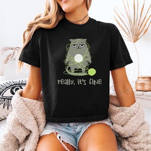 May include: Black t-shirt featuring a grumpy gray cat tangled in yarn with a green ball of yarn. The text "really, it's fine" is printed below the cat. The shirt is worn by a person.