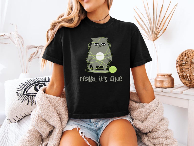May include: Black t-shirt featuring a grumpy gray cat tangled in yarn with a green ball of yarn. The text "really, it's fine" is printed below the cat. The shirt is worn by a person.