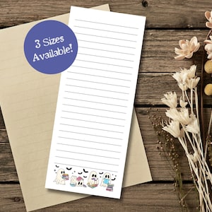 May include: A lined notepad with a decorative border featuring cartoon ghosts reading books, with bats flying above. A blue circle with the text "3 Sizes Available!" is visible. The notepad rests on a beige paper, all on a wooden surface.