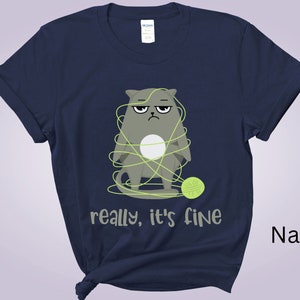May include: Navy blue t-shirt featuring a grumpy gray cat tangled in yarn, with a green ball of yarn nearby. The text "really, it's fine" is printed below the cat. The shirt is a solid navy color.