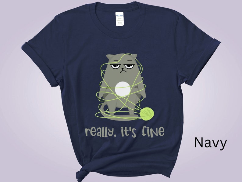 May include: Navy blue t-shirt featuring a grumpy gray cat tangled in yarn, with a green ball of yarn nearby. The text "really, it's fine" is printed below the cat. The shirt is a solid navy color.