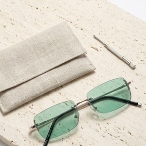 May include: Rimless rectangular sunglasses with green tinted lenses and silver frames. A small silver screwdriver and a beige linen glasses case are also present. The glasses case is folded over.