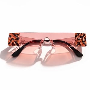 May include: Rimless sunglasses with a pink tint and black arms. The temples feature a brown and orange snakeskin pattern. The sunglasses are set against a white background, casting a shadow.