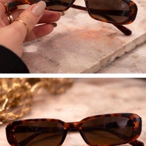 May include: Rectangular sunglasses with a tortoiseshell pattern. The frames are brown with dark lenses. The sunglasses are shown on a textured surface.