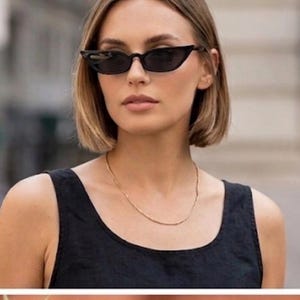 May include: A woman wearing black cat-eye sunglasses and a gold necklace. She is wearing a black tank top. The background is a blurred city street with a coffee shop.