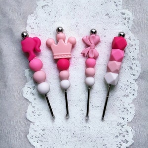 May include: Four silicone teethers with different shapes and colors. The teethers are pink, white, and light pink. Each teether is attached to a silver metal stick.