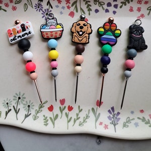 May include: Five colorful, beaded, metal skewers with different animal and cat-themed charms. The charms include a cat in a basket, a golden retriever, a rainbow paw print, and a black pug. The skewers are on a white plate with a floral design.