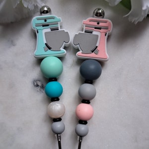 May include: Two silicone beaded necklaces with a miniature kitchen mixer charm at the top. One necklace has teal, turquoise, white, and gray beads. The other necklace has pink, gray, and white beads.