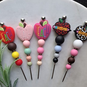 May include: Five colorful apple-shaped silicone toppers with the word "Teach" on them. Each topper is attached to a metal skewer with colorful beads. The toppers are perfect for decorating drinks or food.