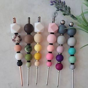 May include: Six colorful silicone bead and metal tipped tools. The tools are arranged in a row from left to right. The beads are in various colors, including white, brown, beige, pink, purple, and turquoise.
