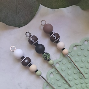 May include: Three different colored stirring sticks with a football design. Each stick has a white football with black stitching and a black bead, followed by a bead in a different color: brown, black, and green. The sticks are on a green silicone mat with small circles.