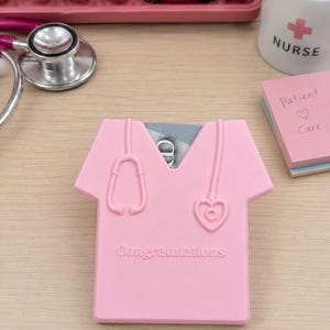 Op de afbeelding: Een roze kaarthouder in de vorm van een medische scrubtop, met een stethoscoopontwerp en het woord "Congratulations". Het ligt op een houten bureau, met een toetsenbord, een mok met "Nurse" en plaknotities op de achtergrond.