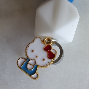 May include: A white, gold, and red Hello Kitty charm with a blue and white outfit. The charm is attached to a silver ring.