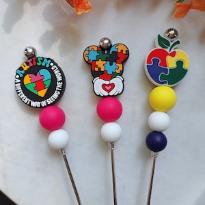 May include: Three colorful silicone charms with puzzle pieces and the words "Autism, A Different Way of Seeing the World" on a white background. Each charm is attached to a silver skewer.