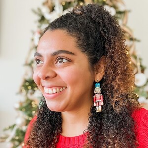 Pu&ograve; includere: Una donna con lunghi capelli castani ricci indossa un maglione rosso e orecchini a forma di schiaccianoci. Gli orecchini sono in legno e dipinti con colori vivaci.