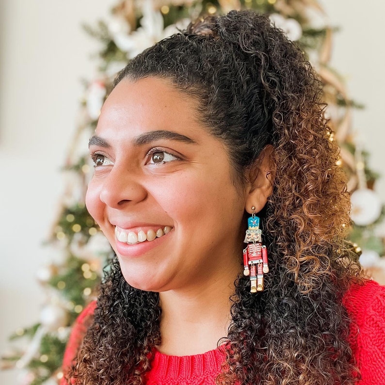 Pu&ograve; includere: Una donna con lunghi capelli castani ricci indossa un maglione rosso e orecchini a forma di schiaccianoci. Gli orecchini sono in legno e dipinti con colori vivaci.