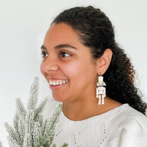 Pu&ograve; includere: Una donna indossa un maglione bianco con un paio di orecchini a forma di schiaccianoci bianchi e dorati. Gli orecchini sono pendenti e presentano una statuetta di schiaccianoci bianca con dettagli dorati.