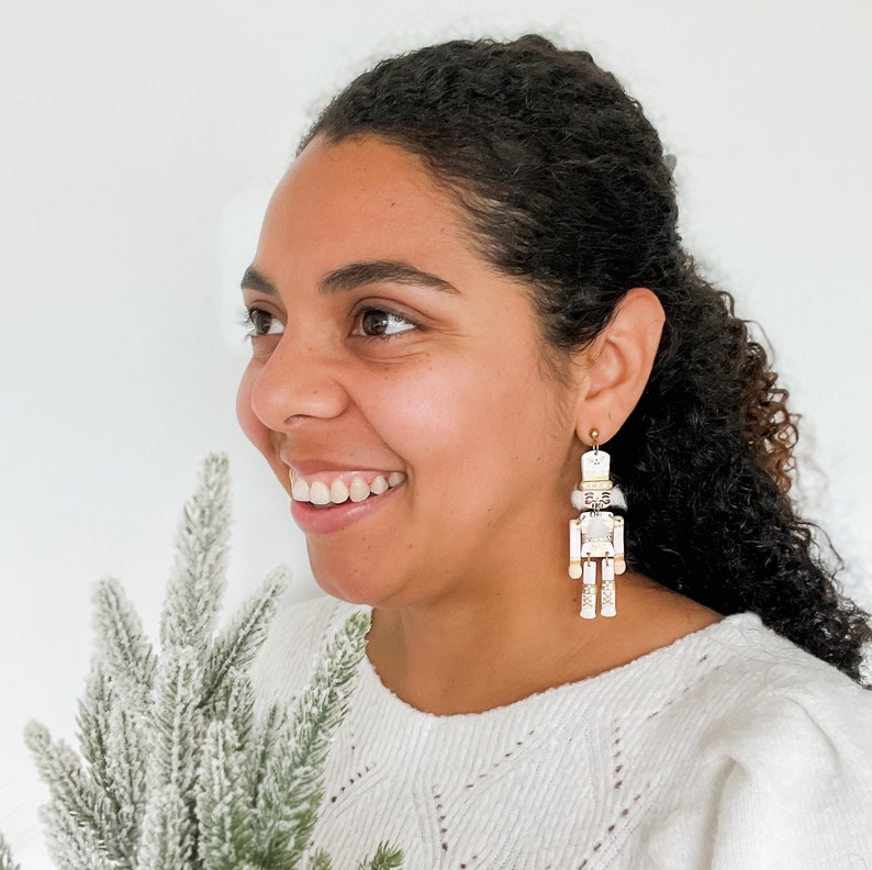 Pu&ograve; includere: Una donna indossa un maglione bianco con un paio di orecchini a forma di schiaccianoci bianchi e dorati. Gli orecchini sono pendenti e presentano una statuetta di schiaccianoci bianca con dettagli dorati.