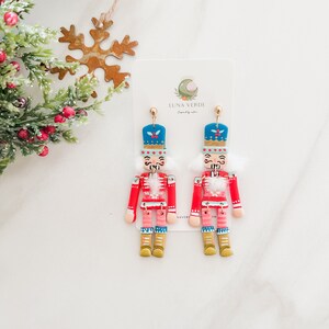 Pu&ograve; includere: Un paio di orecchini a tema natalizio con un design a forma di schiaccianoci. Gli orecchini sono realizzati in pasta polimerica e dipinti in rosso, blu, bianco e oro. Le figure di schiaccianoci indossano un cappotto rosso, un cappello blu e un bordo di pelliccia bianca. Gli orecchini sono appesi a ganci dorati.
