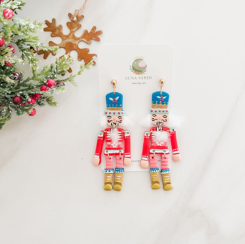 Pu&ograve; includere: Un paio di orecchini a tema natalizio con un design a forma di schiaccianoci. Gli orecchini sono realizzati in pasta polimerica e dipinti in rosso, blu, bianco e oro. Le figure di schiaccianoci indossano un cappotto rosso, un cappello blu e un bordo di pelliccia bianca. Gli orecchini sono appesi a ganci dorati.