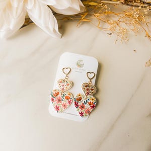 以下が含まれることがあります： 花柄の金色のハート型のイヤリングのペア。イヤリングは重ねてあり、小さなハートが大きなハートの上にあります。花柄はピンク、赤、オレンジ、黄色の色合いです。
