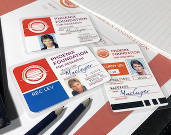 May include: Three plastic ID cards with a red, white, and blue logo of a bird in flight. The cards have the text "Phoenix Foundation for Research" and the name "MacGyver" printed on them. The cards have a photo of a man with brown hair and a signature.