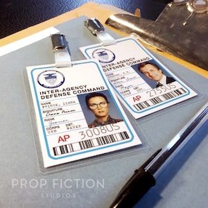 Puede incluir: Dos tarjetas de identificación de plástico blanco con texto negro y códigos de barras. La tarjeta superior tiene el nombre "Trevor, S.R." y el rango "Major". La tarjeta inferior tiene el nombre "Prince, Diana" y el rango "Yeoman".