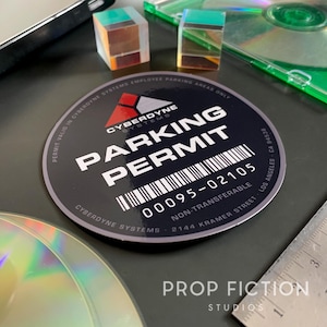 Könnte beinhalten: Ein runder, grau-schwarzer Parkscheinaufkleber mit dem Text "CYBERDYNE SYSTEMS PARKING PERMIT" und der Nummer "00095-02105". Der Aufkleber enthält auch den Text "NICHT ÜBERTRAGBAR" und die Adresse "2144 KRAMER STREET LOS ANGELES CA 94538".