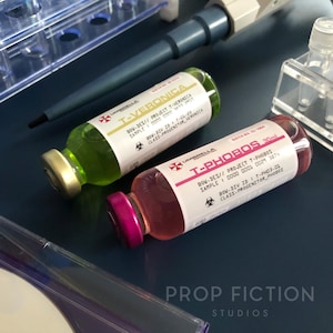 May include: Two laboratory vials, one with green liquid and the other with pink liquid, labelled "T-VERONICA" and "T-PHOBOS". The vials have white labels with black text, including "Umbrella" branding and biohazard symbols. The background includes laboratory equipment.