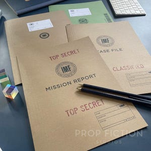 May include: A collection of brown and green file folders with the text "TOP SECRET", "MISSION REPORT", and "CASE FILE". The folders have "IMF" logos and are accompanied by two black pencils.