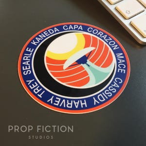 May include: A round sticker with a retro design featuring a stylized sun, a white and blue object, and the text "SEARLE KANEDA CAPA CORAZON MACE CASSIDY HARVEY TREY" in a blue border. The sticker also has the text "PROP FICTION STUDIOS".