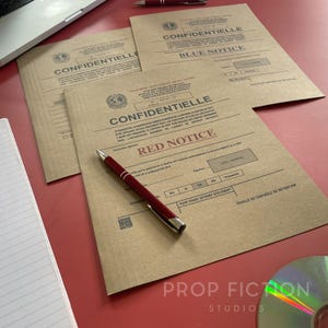 Puede incluir: Tres carpetas de manila de color tostado con texto impreso en negro que incluye las palabras CONFIDENTIELLE, RED NOTICE y BLUE NOTICE. Un bolígrafo rojo y plateado descansa sobre la carpeta superior. Un cuaderno rayado y un CD son visibles.