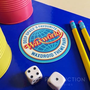 Puede incluir: Una pegatina redonda con el texto "DEEP SPACE'S FAVOURITE Waxworld WAXDROID THEME PARK" en rojo, amarillo y azul. Dos lápices amarillos con gomas rosas y dos dados blancos con puntos negros están sobre una superficie azul.