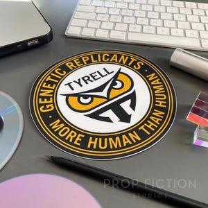 May include: A round sticker with the text "GENETIC REPLICANTS - MORE HUMAN THAN HUMAN" in a yellow and black circular design. The sticker features an owl-like graphic with the name "TYRELL" in the center. The sticker is on a desk with other objects.