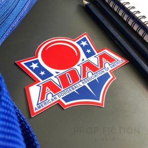 May include: A sticker featuring the ADAA logo, the American Dodgeball Association of America. The logo has a red circle over the word ADAA in white, with blue accents and white stars. The sticker is on a dark surface with pencils and a notebook.