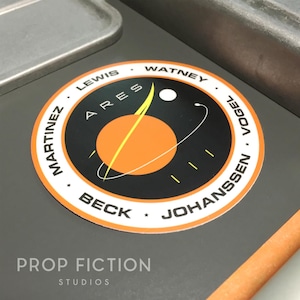 May include: A circular sticker with a black and orange space-themed design. The sticker features the word "ARES" in white, surrounded by names and a graphic of a planet with a yellow line. The text "PROP FICTION STUDIOS" is at the bottom.