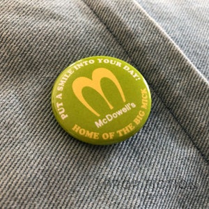 Könnte beinhalten: Ein grüner und gelber Knopf mit dem Text "Put a smile into your day! McDowell's Home of the Big Mick"