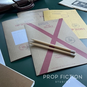 Puede incluir: Una colección de documentos de estilo vintage con las etiquetas "Top Secret" y "Operation Mincemeat" con detalles en rojo. Dos lápices descansan sobre los documentos. La imagen incluye un maletín de cuero y el texto "Prop Fiction Studios."