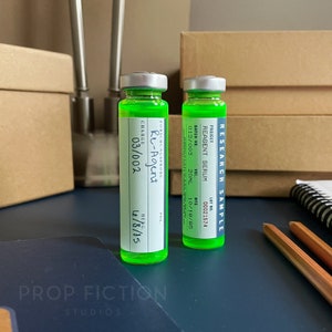 Peut inclure: Deux petites bouteilles en verre contenant un liquide vert et des étiquettes blanches. Les étiquettes portent le texte suivant : "RESEARCH SAMPLE", "PROJECT", "REAGENT SERUM", "BATCH NO. 012/003", "LOT NO. 00021574", "VOL. 20ML", "MFD 10/18/85", "FORSCHUNGSPROBE", "Re-Agent", "CHARGE 03/002", "VOL.", "HERG. 6/8/85".