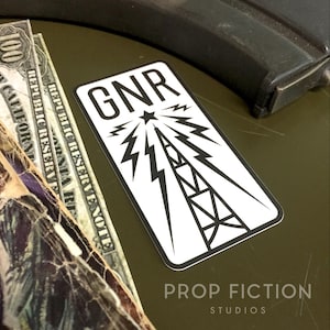 May include: A white rectangular sticker with the letters "GNR" at the top, a star, and a stylized radio tower design. The sticker is on a green surface, with US currency and a dark object in the background. The text "PROP FICTION STUDIOS" is at the bottom.