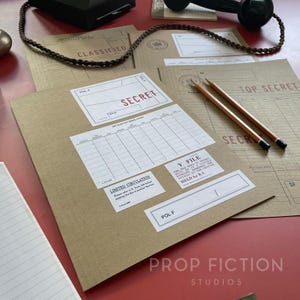 以下が含まれることがあります： SECRET、TOP SECRET、CLASSIFIED、Y FILE、LIMITED CIRCULATIONなどの文字が印刷されたラベルが付いた茶色の紙製フォルダーのコレクション。フォルダーの上には木製の鉛筆が2本置かれ、編み込まれたコードが黒い物体につながっている。