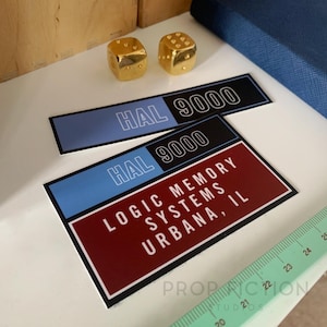 Op de afbeelding: Twee gouden dobbelstenen en twee rechthoekige stickers. De stickers hebben een blauw en zwart gedeelte met de tekst "HAL 9000". Eén sticker heeft ook de tekst "LOGIC MEMORY SYSTEMS URBANA, IL".