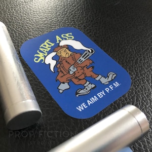 May include: A blue rectangular sticker with the words "SMART ASS" in yellow and a cartoon character holding a gun. The text "WE AIM BY P.F.M." is below the character. Two silver cylindrical objects are in the foreground.