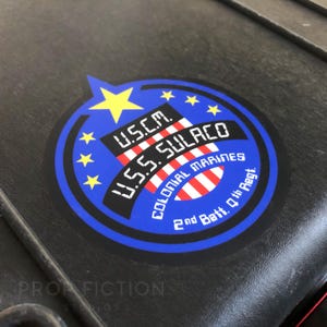 Puede incluir: Una pegatina circular con un borde azul y una estrella amarilla en la parte superior. La pegatina presenta el texto "U.S.C.M. U.S.S. SULACO COLONIAL MARINES 2nd Batt. 7th Regt." en letras blancas. La pegatina está sobre una superficie negra.
