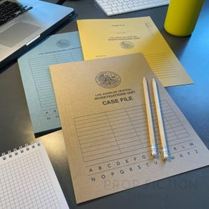 May include: Three case files from the Los Angeles Central Investigations Unit are displayed, with the text "CASE FILE" and the unit's emblem. Two pencils rest on the brown file. A notebook, laptop, and keyboard are also visible.